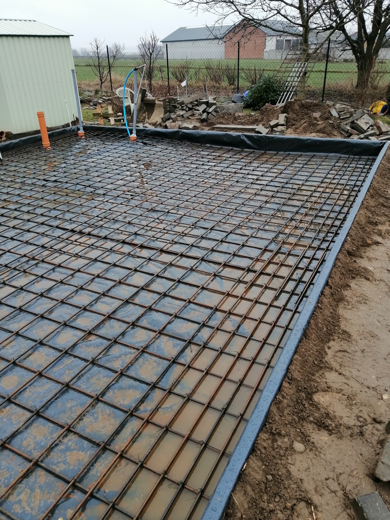 Przygotowany teren pod wylewkę z ułożoną siatką zbrojeniową, folią i rurami instalacyjnymi, widoczne elementy szalunku, w tle budynki gospodarcze i resztki kostki brukowej.