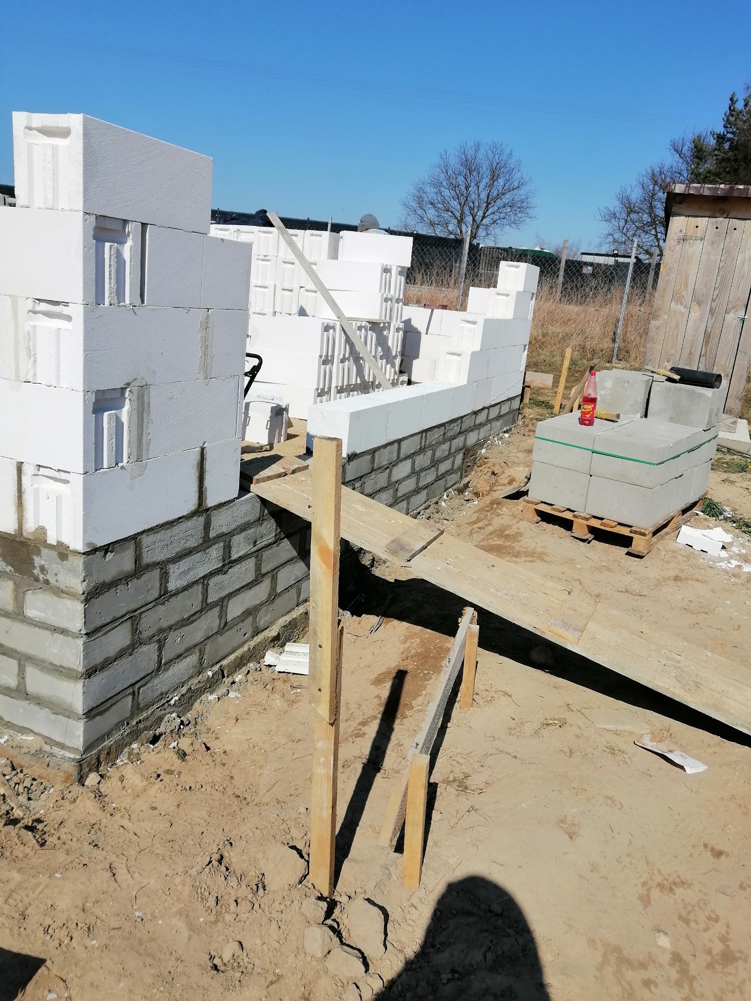 Wznoszenie ścian budynku z białych bloczków na fundamencie z szarej cegły, widoczne drewniane podpory i paleta z materiałami budowlanymi w tle, teren budowy w słoneczny dzień.