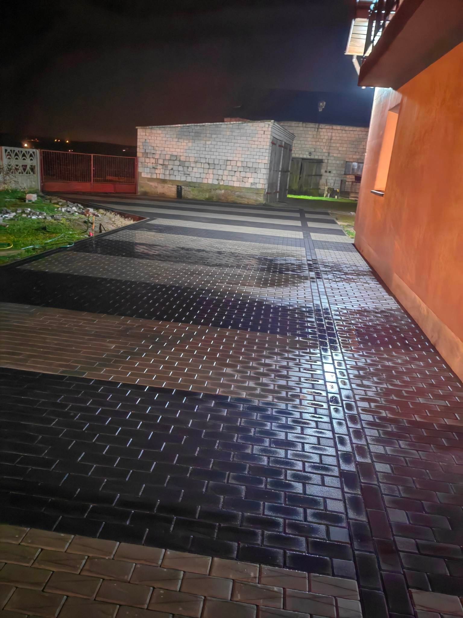 Świeżo ułożona, mokra kostka brukowa w odcieniach brązu i czerni, tworząca wzór pasów, widoczna w nocy przy oświetleniu zewnętrznym, z budynkiem w tle.