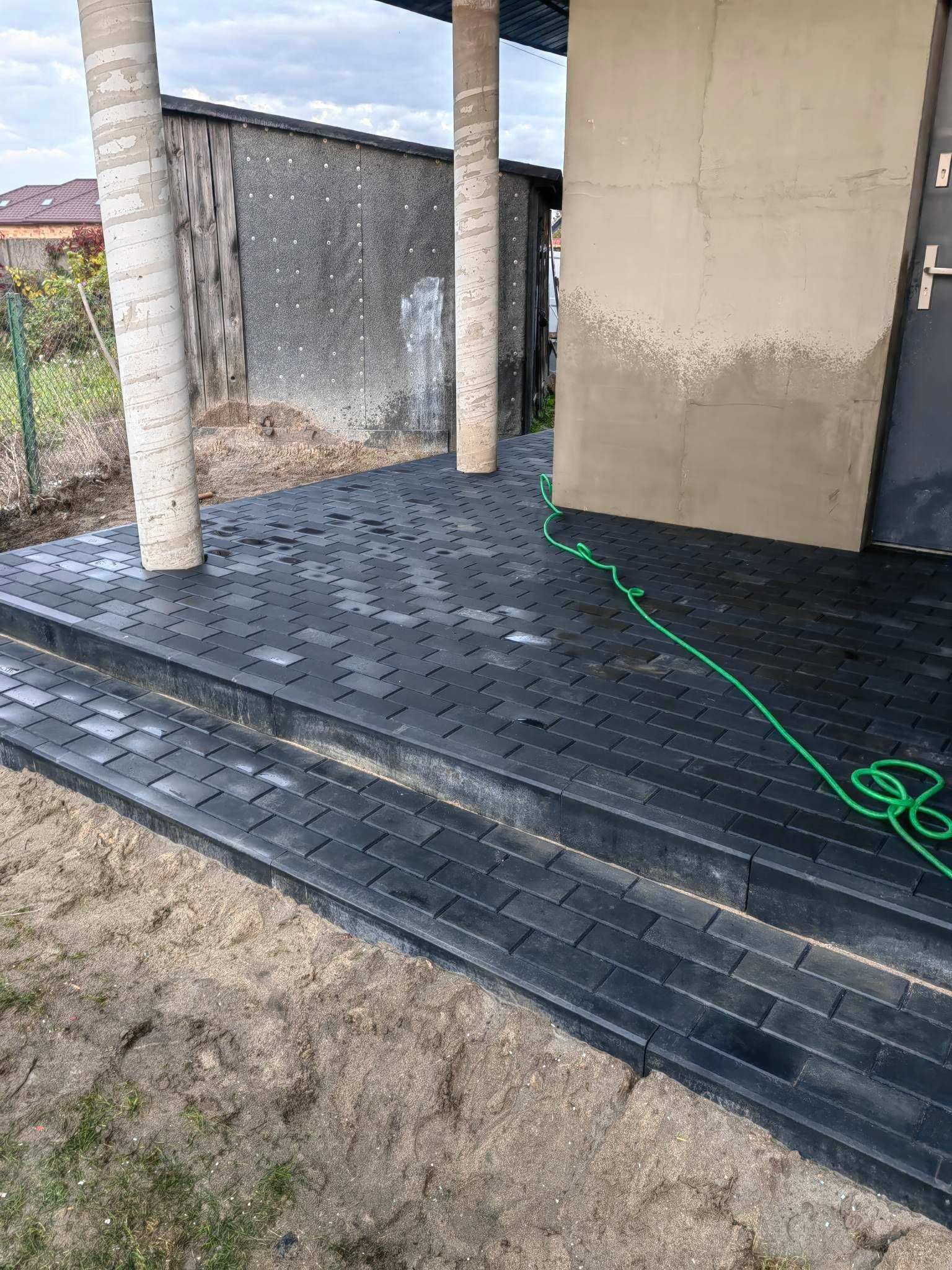 Nowo ułożona, czarna kostka brukowa tworząca schody wejściowe do budynku, z widocznymi kolumnami i zielonym wężem ogrodowym na powierzchni.