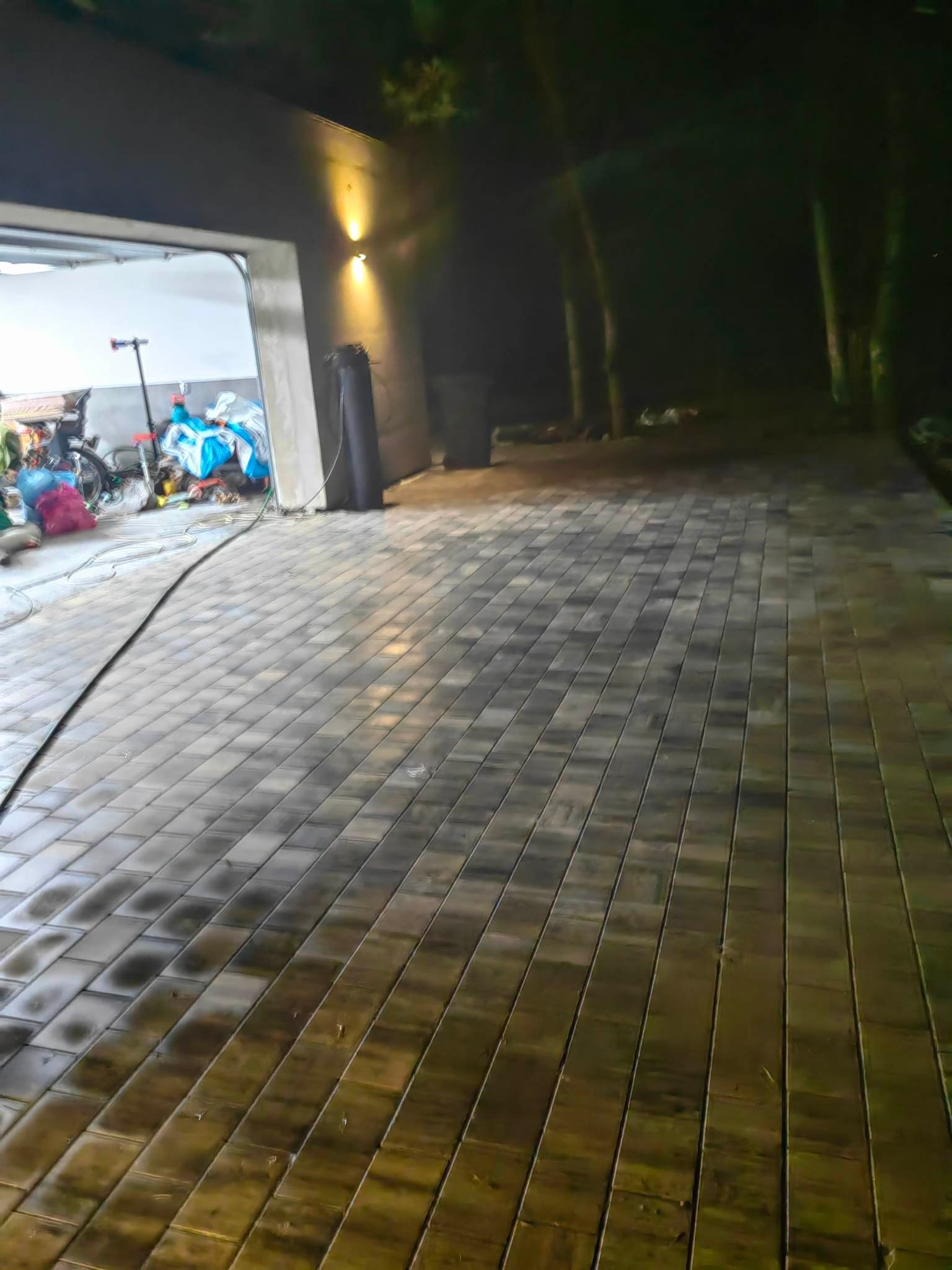 Świeżo ułożona, mokra kostka brukowa w odcieniach szarości i brązu, prowadząca do otwartego garażu z widocznym bałaganem wewnątrz, oświetlona dwoma lampami ściennymi i światłem z garażu, w tle...