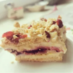 Kawałek warstwowego ciasta z kremem, kruszonką i czerwonymi owocami na białym talerzu, sfotografowany w naturalnym świetle.