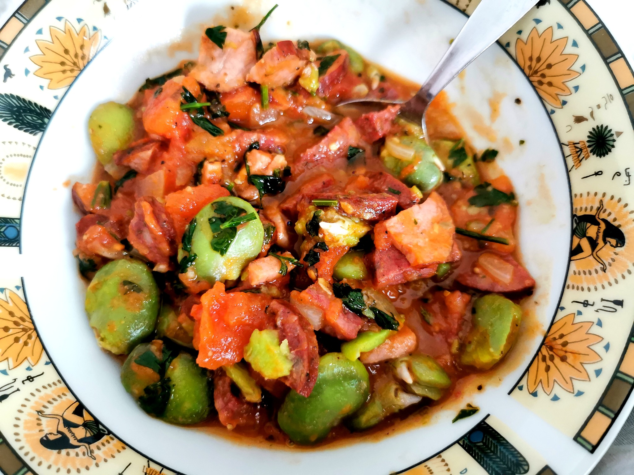 Gulasz z bobu, pomidorów i wędzonki w białym talerzu z dekoracyjnym brzegiem, widoczna łyżka