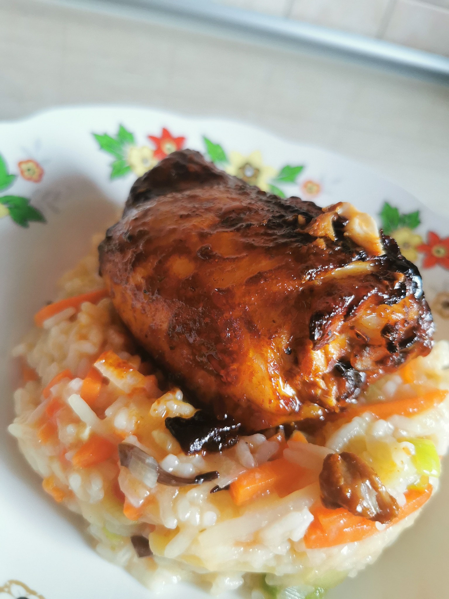 Pieczona noga kurczaka z błyszczącą skórką, ułożona na risotto z marchewką i grzybami, podana na talerzu z kwiatowym wzorem.