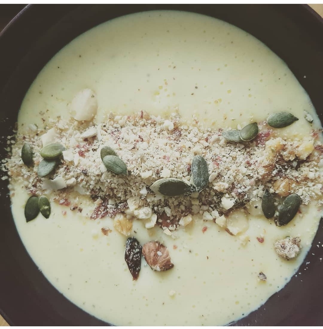 Kremowa zupa w ciemnej misce, posypana mieszanką orzechów, nasion dyni i okruchów, ułożona w centralnym pasie.