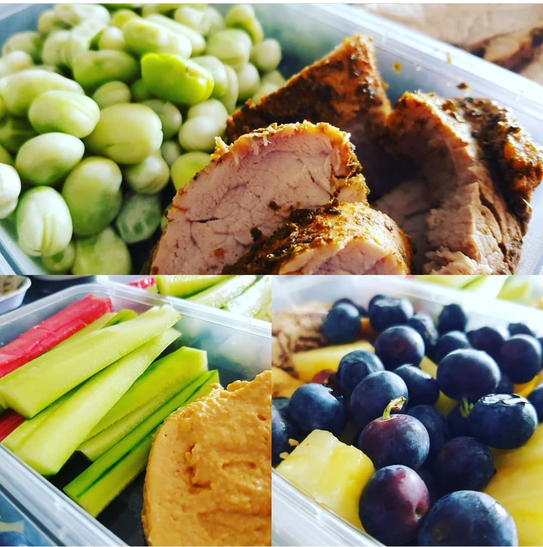 Lunchbox z różnorodnymi składnikami: fasola, pieczona wieprzowina z przyprawami, słupki ogórka i papryki, hummus oraz kostki ananasa z borówkami.