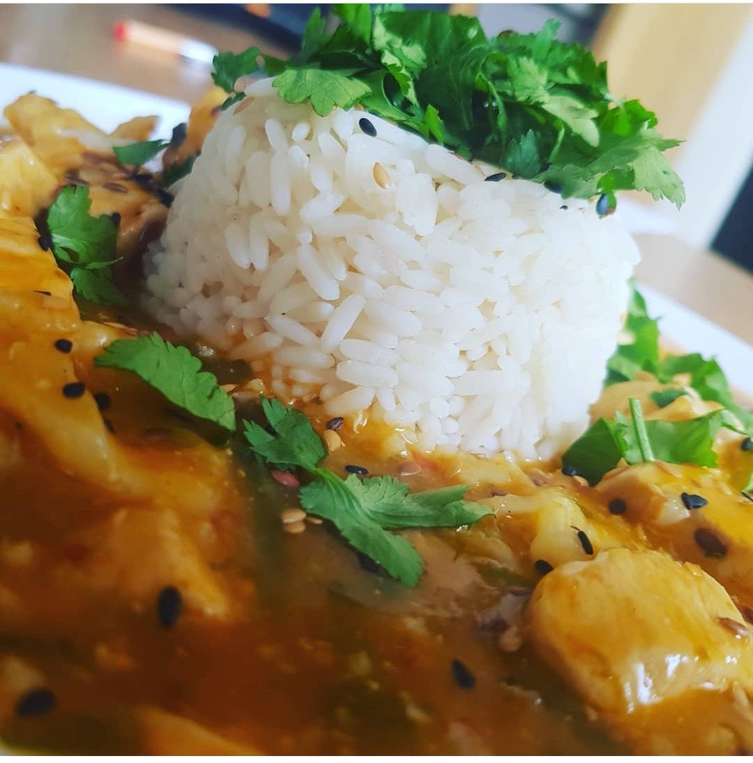 Porcja ryżu uformowana w kopiec, polana sosem z kawałkami kurczaka i posypana świeżą kolendrą oraz czarnym sezamem, podana na białym talerzu.