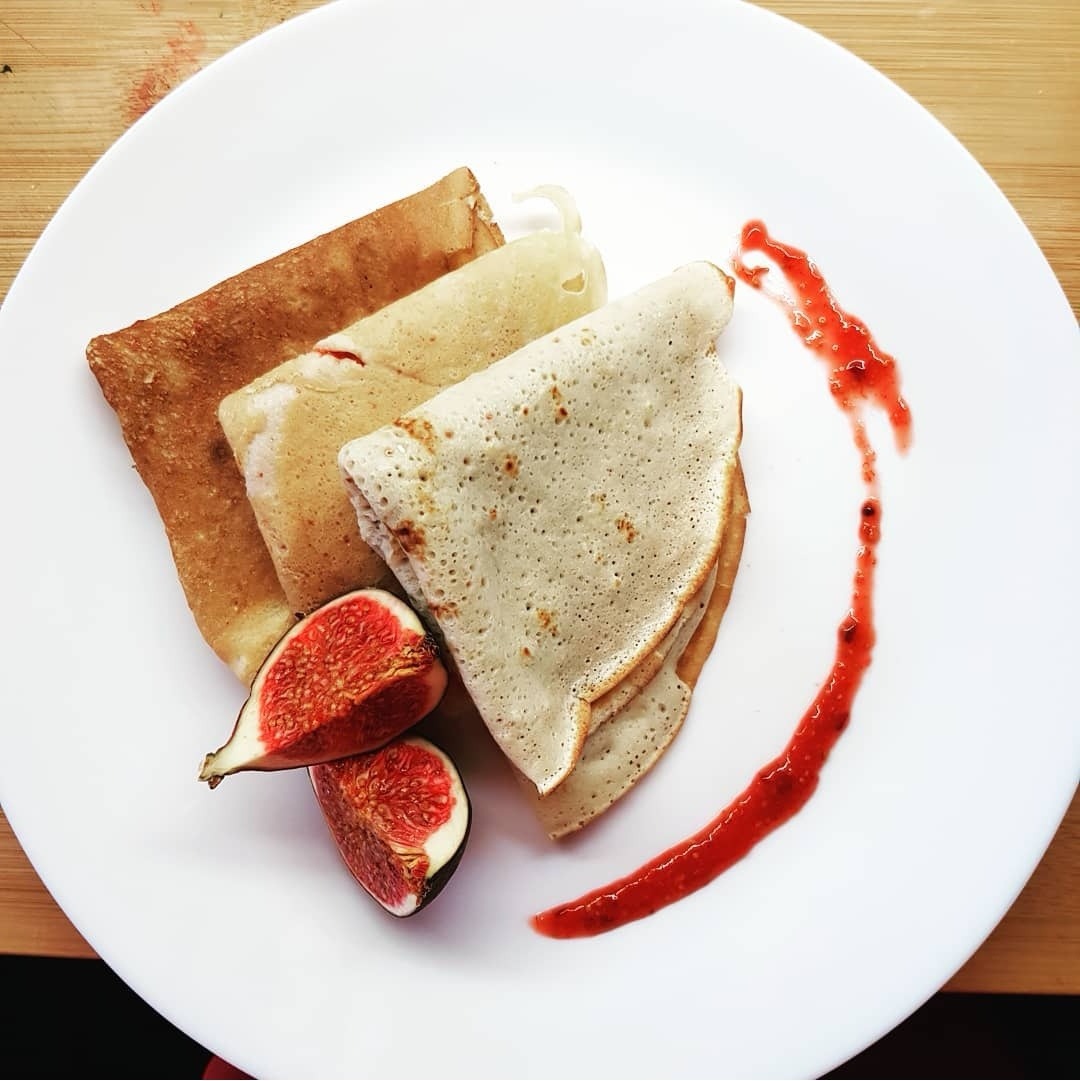 Trzy naleśniki złożone w trójkąty o różnym stopniu wysmażenia, podane na białym talerzu z połówkami figi i dekoracją z czerwonego sosu.