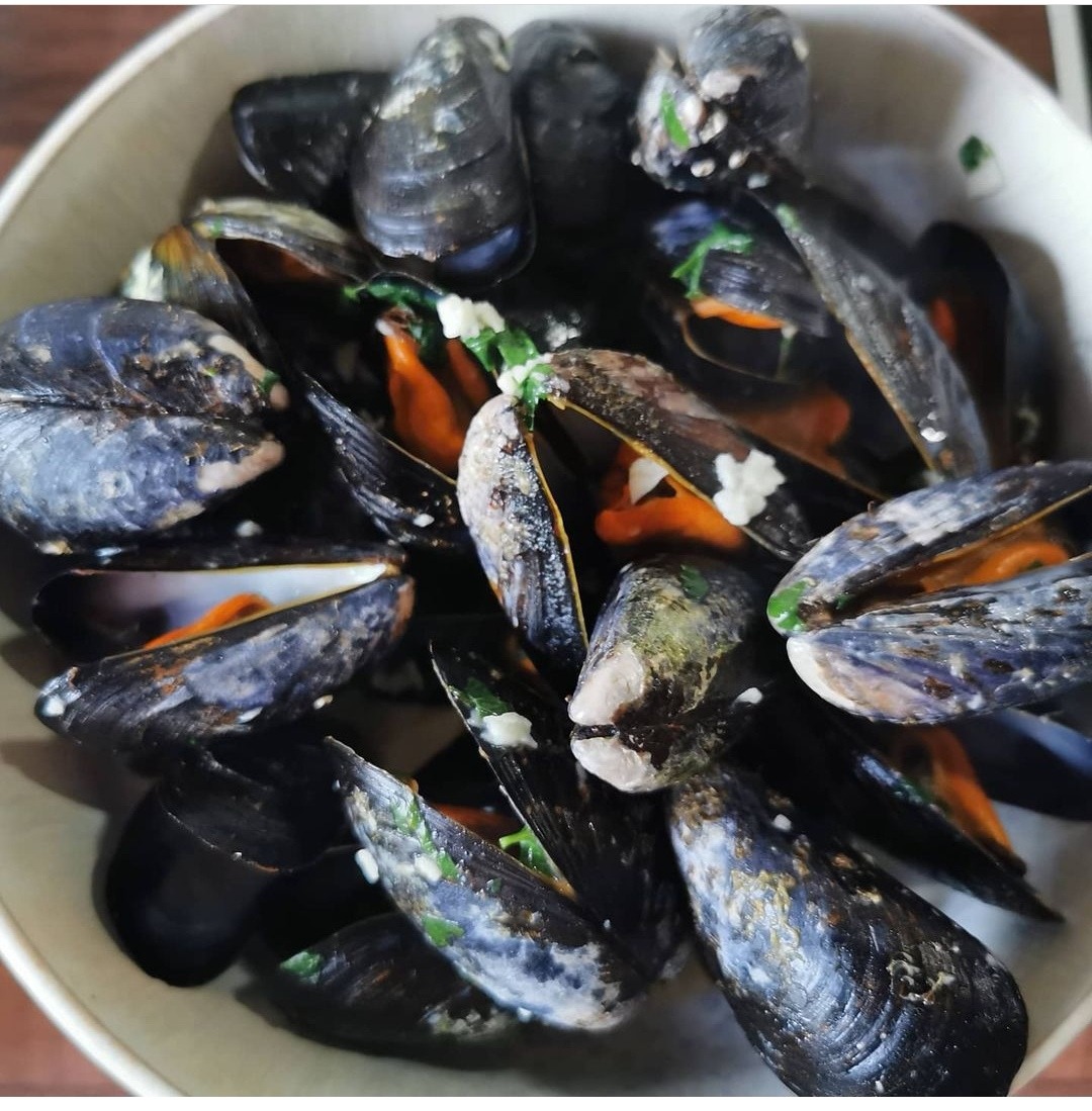 Porcja gotowanych muli w muszlach, podanych w jasnej misce, udekorowanych ziołami i sosem czosnkowym.