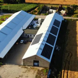 Widok z lotu ptaka na dwa duże budynki gospodarcze z panelami słonecznymi na dachu jednego z nich, otoczone polami i zabudowaniami gospodarczymi.