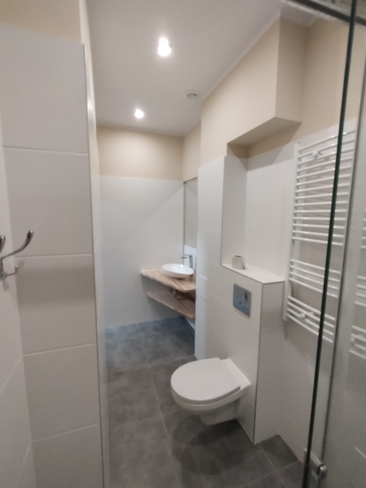 Nowoczesna łazienka z umywalką na drewnianym blacie, podwieszaną toaletą i grzejnikiem drabinkowym, widok zza szklanej kabiny prysznicowej.