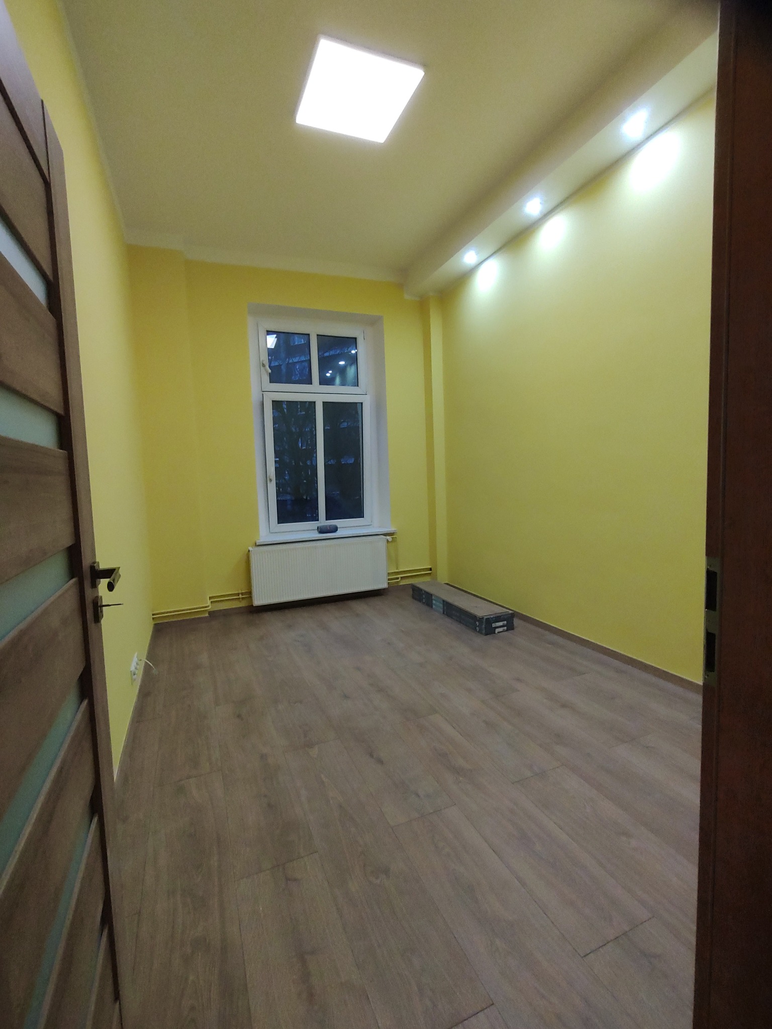 Pusty pokój z żółtymi ścianami, panelami podłogowymi, białym grzejnikiem pod oknem i listwą oświetleniową LED pod sufitem. Na podłodze leżą paczki paneli.