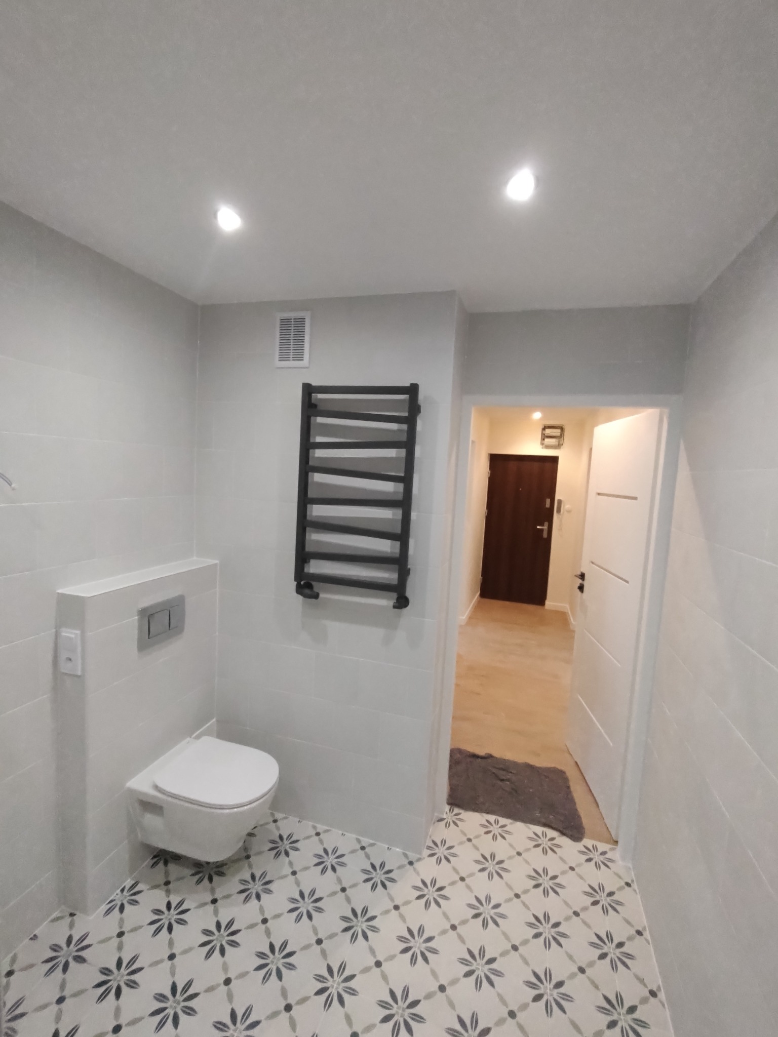 Nowoczesna łazienka w jasnych odcieniach szarości z podwieszaną toaletą, grzejnikiem drabinkowym w kolorze czarnym i wzorzystą podłogą w kwiaty.