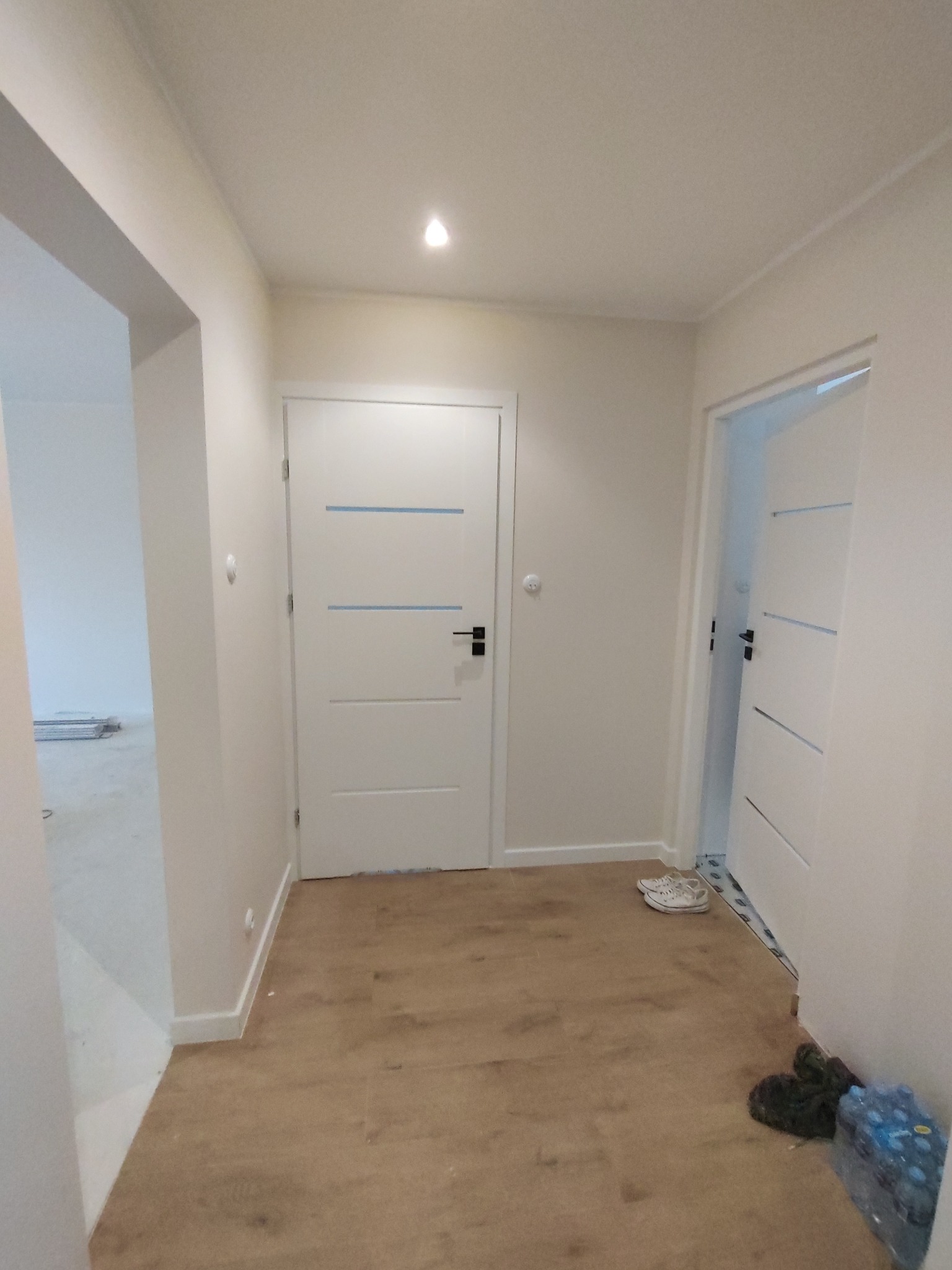 Korytarz w trakcie wykańczania z panelami podłogowymi, dwiema białymi parami drzwi z niebieskimi akcentami i butami oraz butelkami wody przy ścianie.