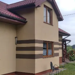 Dom jednorodzinny z beżową elewacją i brązowymi pasami, brązową dachówką, widoczne okna z firankami, krzesło i piłka na trawniku.