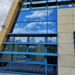 Fasada budynku biurowego z niebieskimi ramami okiennymi, w kt&oacute;rych odbija się błękitne niebo z chmurami i żuraw budowlany, z widoczną kamerą monitoringu na elewacji.