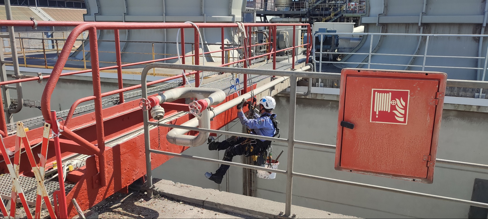 Pracownik w uprzęży, na wysokości, przy rurociągu przemysłowym, z wiadrem farby, maluje rury na tle instalacji przemysłowej i czerwonej konstrukcji stalowej.