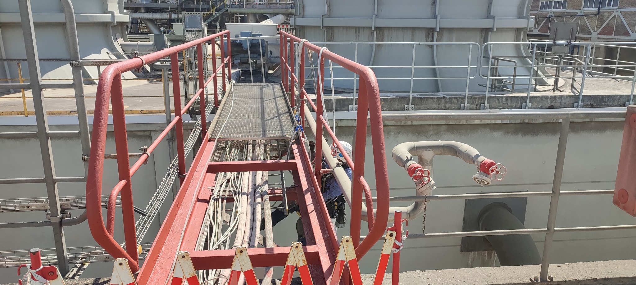 Pracownik w uprzęży na metalowej konstrukcji z czerwonymi barierkami, widoczne rury i elementy instalacji przemysłowej w tle.