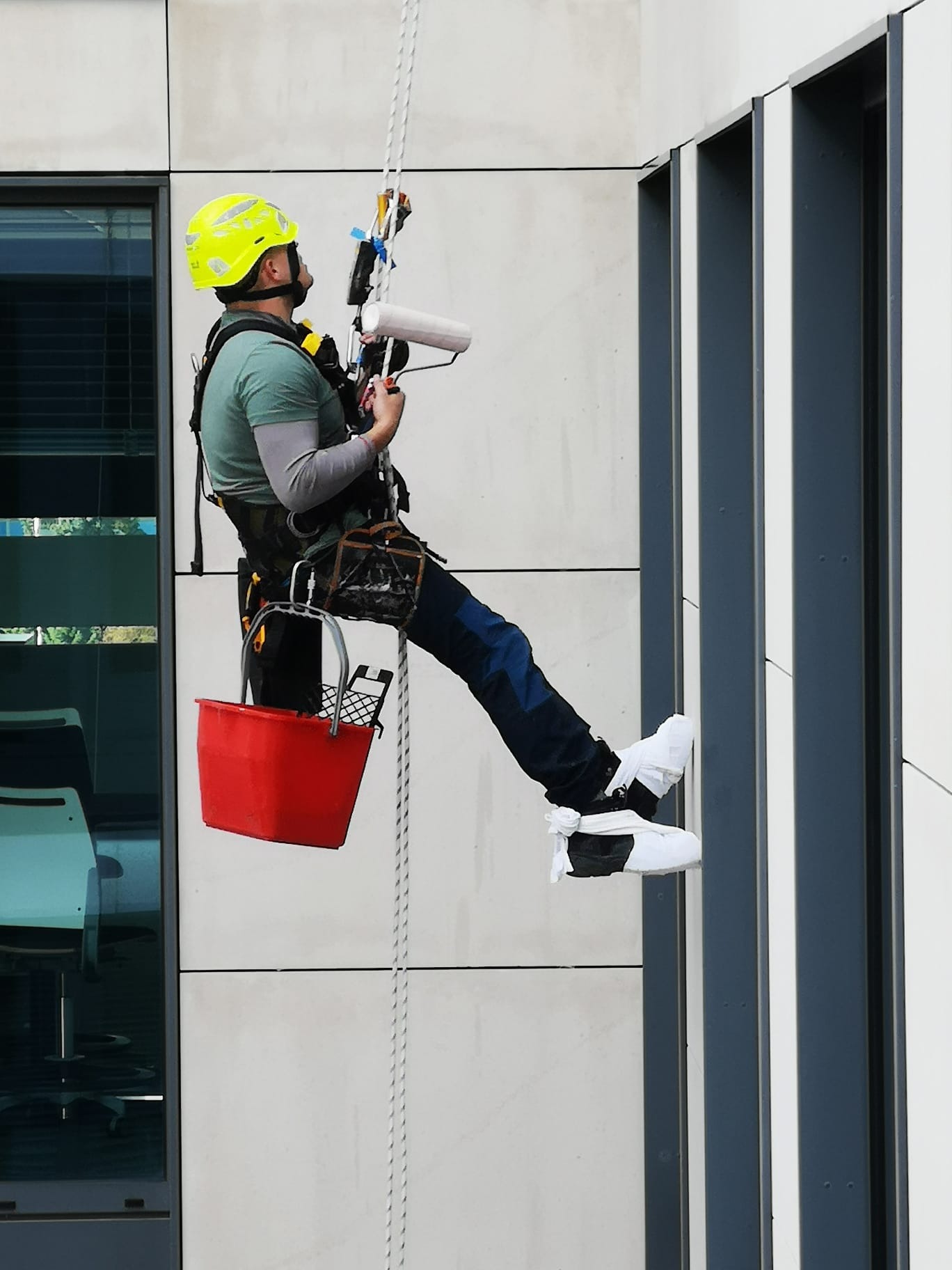 Pracownik w uprzęży, z żółtym kaskiem i czerwonym wiadrem, zawieszony na linie przy nowoczesnym budynku, przygotowuje się do pracy z wałkiem malarskim, zabezpieczając buty białymi ochraniaczami.