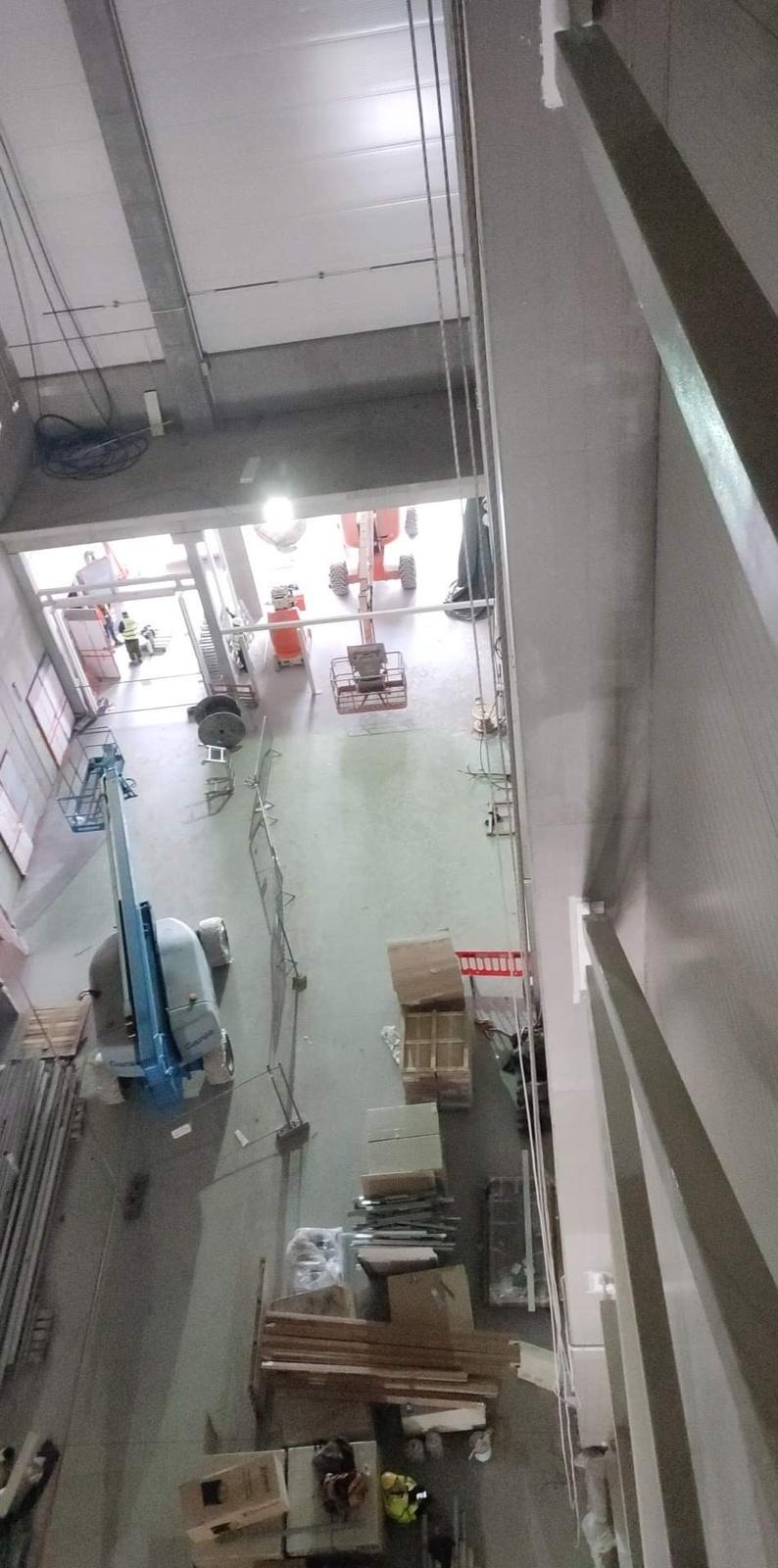 Wysoki magazyn z widocznymi podnośnikami koszowymi, materiałami budowlanymi i pracownikami w oddali, sfotografowany z góry z perspektywy prac wysokościowych.