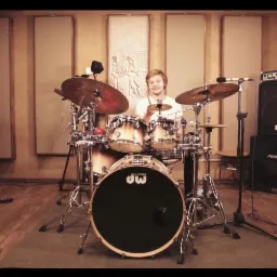 Młody perkusista uśmiecha się siedząc za zestawem perkusyjnym DW w studiu nagraniowym z panelami akustycznymi i wzmacniaczami.