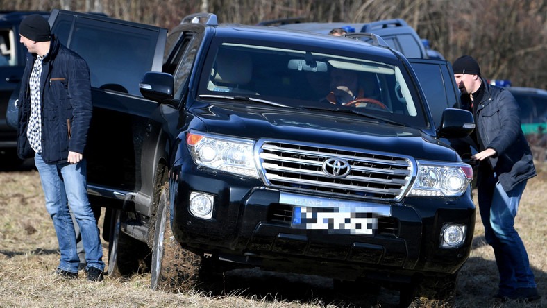 Czarny samochód terenowy Toyota Land Cruiser z otwartymi drzwiami na polnej drodze, dwóch mężczyzn w kurtkach w pobliżu pojazdu.