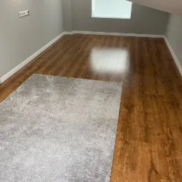 Pokój na poddaszu z ułożonymi panelami podłogowymi w odcieniach brązu i szarym dywanem. Widoczne białe listwy przypodłogowe i szara ściana z gniazdkiem elektrycznym.