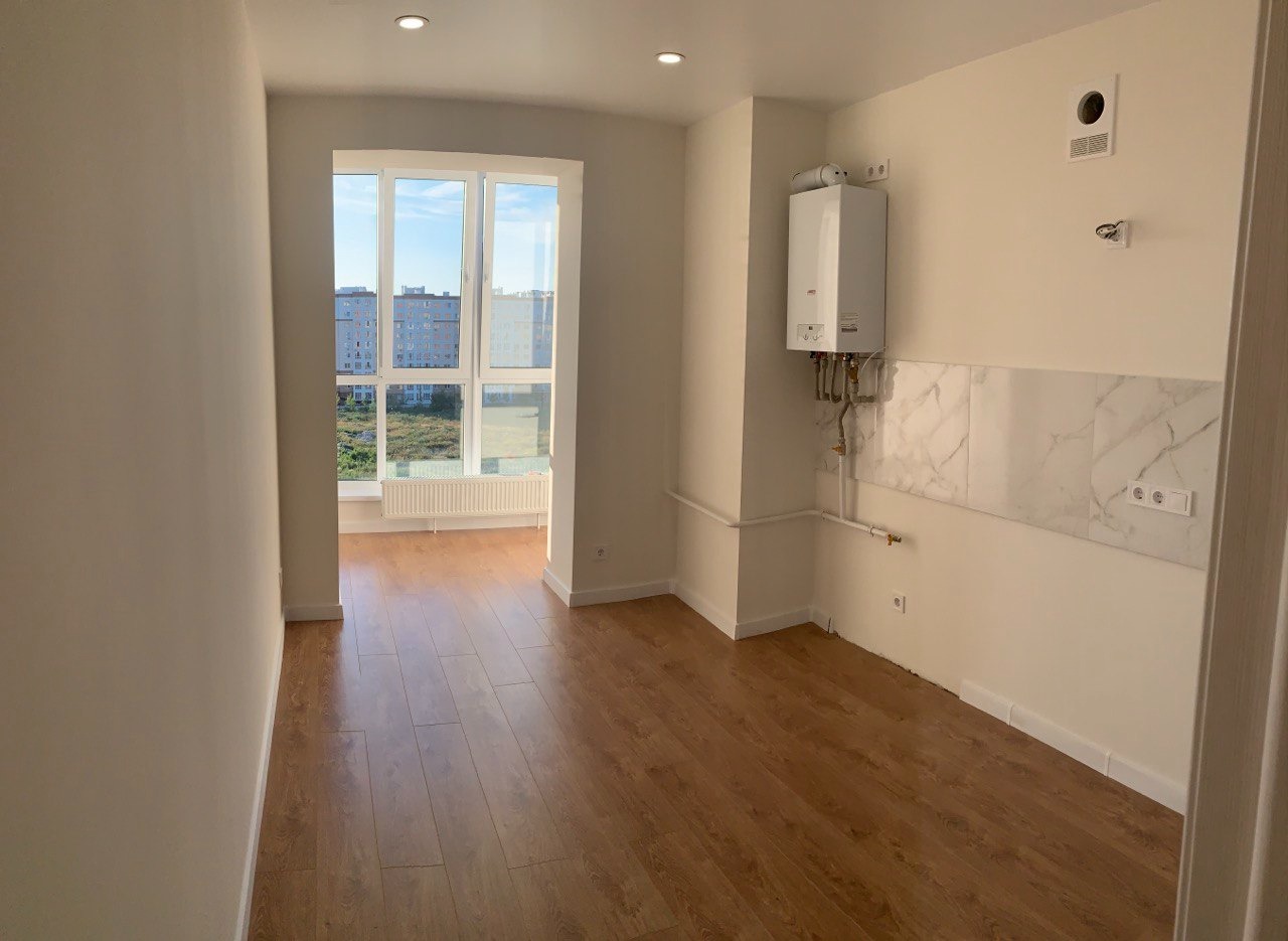 Jasne wnętrze mieszkania po remoncie, z widocznymi panelami podłogowymi, świeżo pomalowanymi ścianami w odcieniach bieli i dużym oknem z widokiem na miasto.