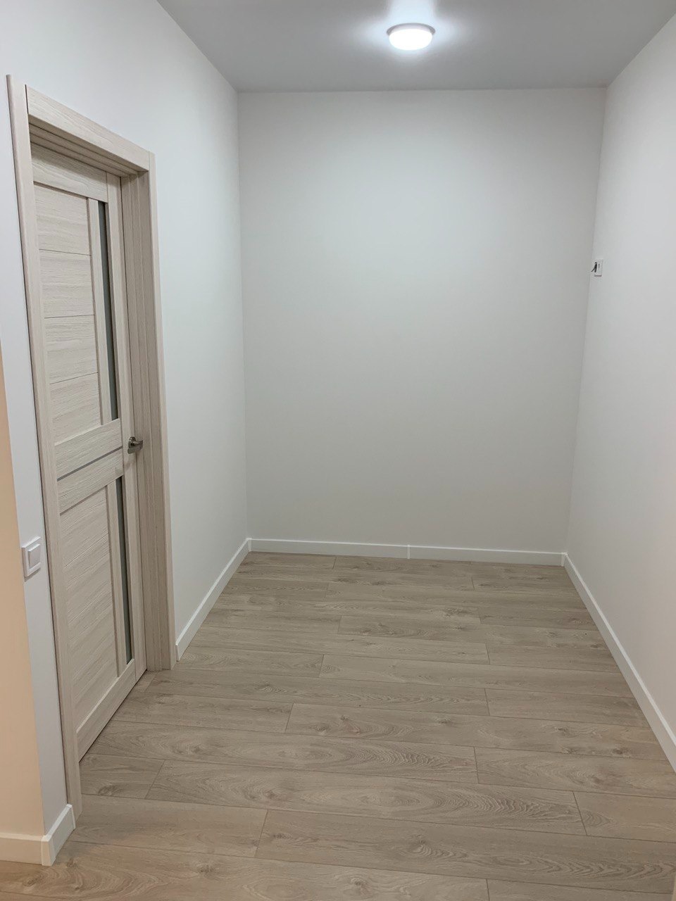 Wąski korytarz z ułożonymi panelami podłogowymi w odcieniach beżu i szarości, świeżo pomalowane ściany na biało, widoczna biała listwa przypodłogowa i drzwi w kolorze paneli.