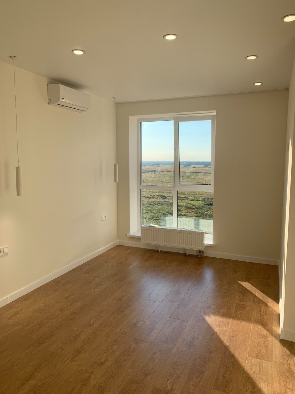 Jasne wnętrze pokoju z drewnopodobnymi panelami podłogowymi, białymi ścianami, grzejnikiem pod oknem i klimatyzatorem na ścianie, oświetlenie punktowe w suficie.