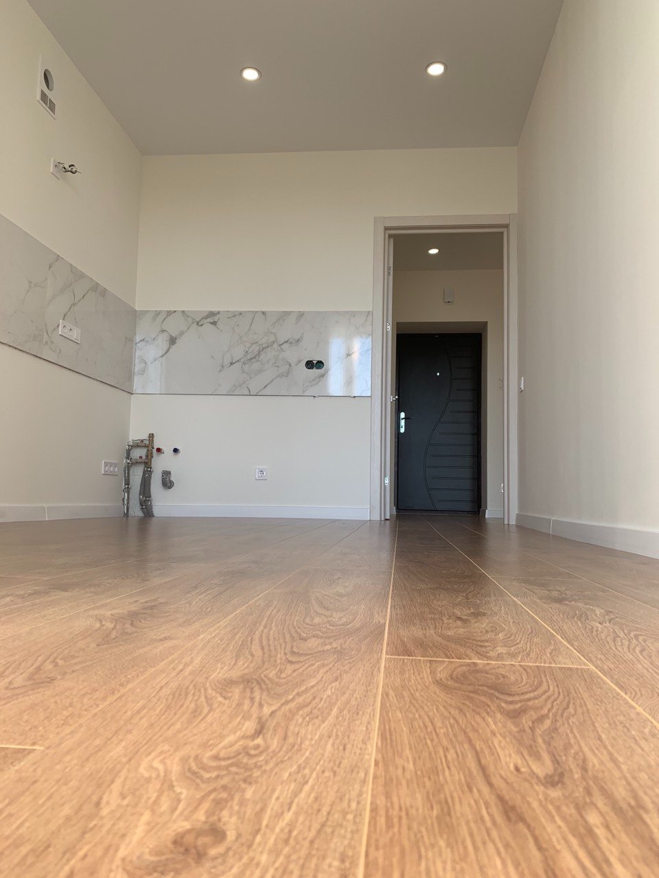 Pusty pokój po remoncie z panelami podłogowymi w odcieniach brązu, widoczne fragmenty instalacji hydraulicznej przy ścianie z marmurową okładziną, neutralne ściany i oświetlenie punktowe.