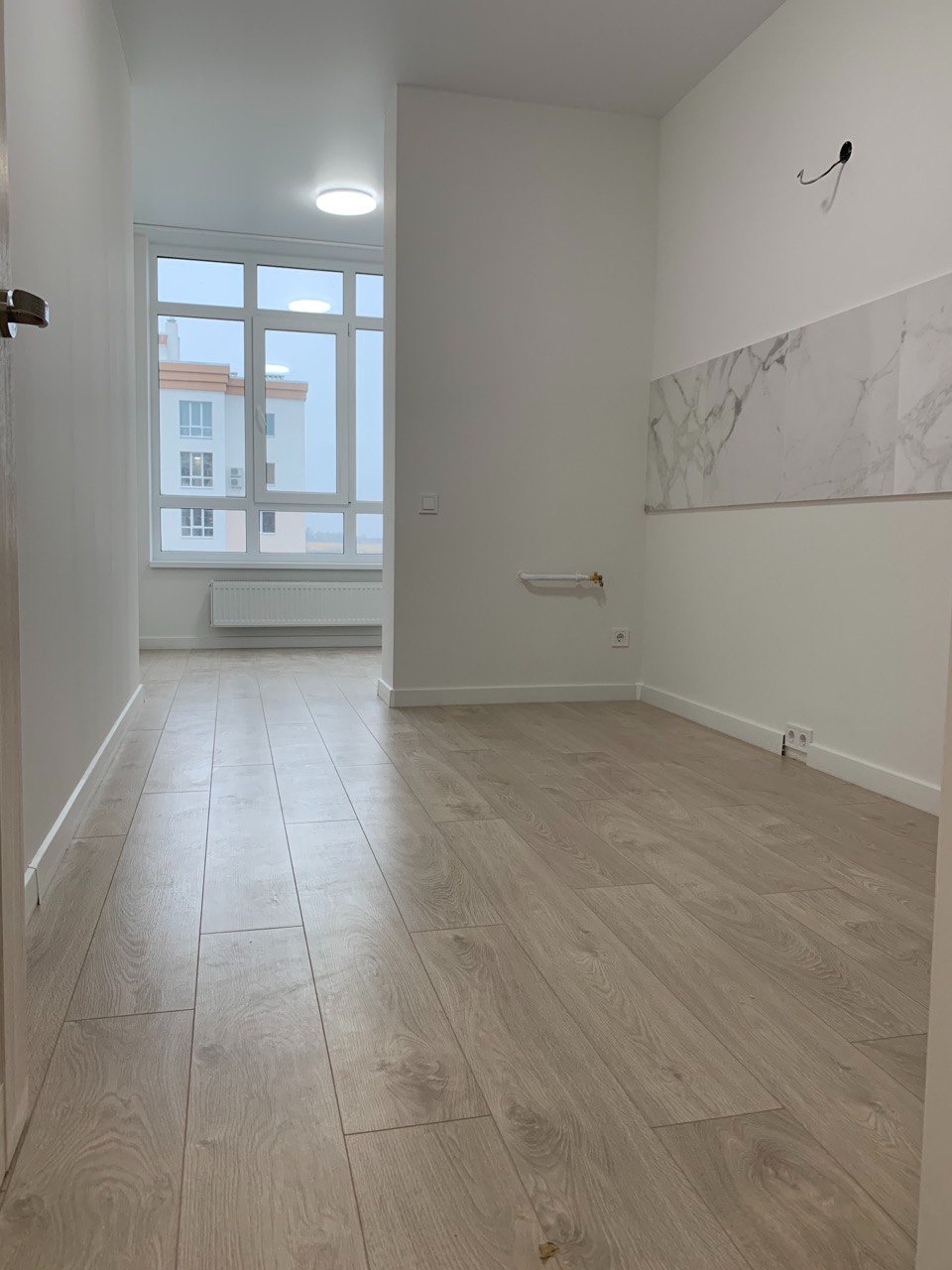 Jasne panele podłogowe w pustym, nowym mieszkaniu z białymi ścianami i fragmentem marmurowej okładziny, widok z korytarza na okno z widokiem na budynek