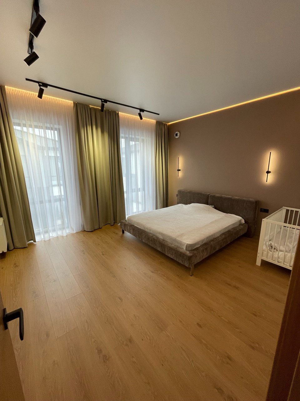 Jasna sypialnia z drewnianą podłogą, beżowymi ścianami, dużym łóżkiem i białym łóżeczkiem dziecięcym, oświetlenie punktowe na suficie.