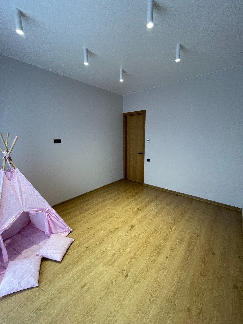 Pokój dziecięcy z jasnymi panelami podłogowymi i różowym tipi, ściany pomalowane na jasny szary kolor, oświetlenie sufitowe w postaci punktowych reflektorów.