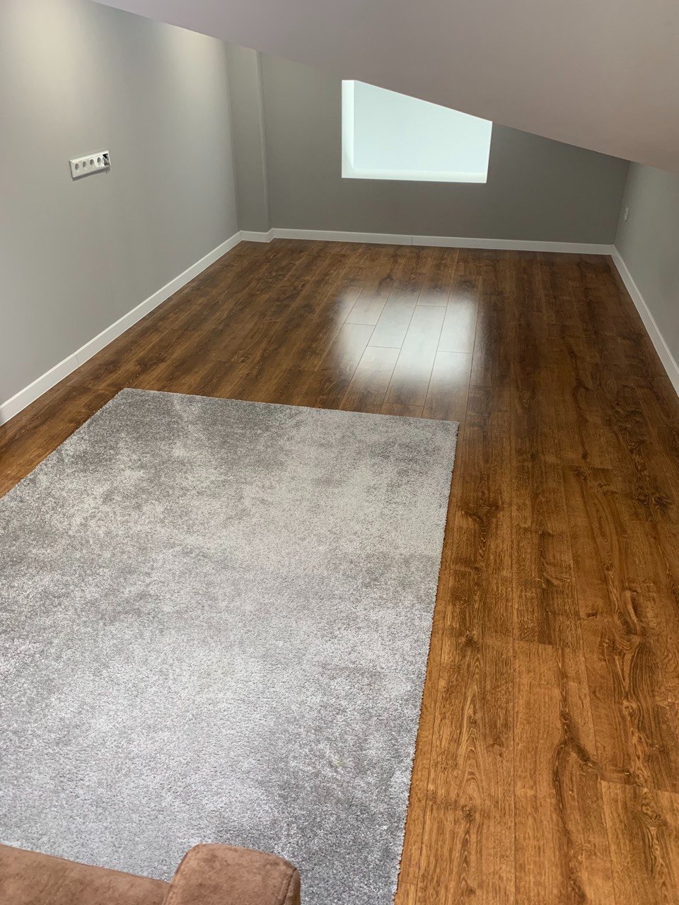 Pokój na poddaszu z ułożonymi panelami podłogowymi w odcieniach brązu i szarym dywanem. Widoczne białe listwy przypodłogowe i szara ściana z gniazdkiem elektrycznym.