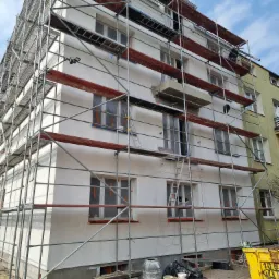 Budynek wielorodzinny w trakcie prac elewacyjnych, okryty rusztowaniami, z widocznymi zabezpieczonymi oknami i przygotowanymi materiałami budowlanymi na ziemi.