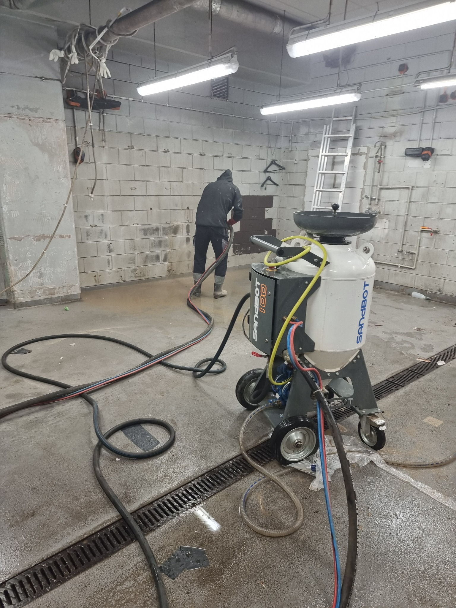 Pracownik w kombinezonie obsługuje urządzenie Sandbot 100 do czyszczenia powierzchni w pomieszczeniu z ceglanymi ścianami i odpływami podłogowymi.