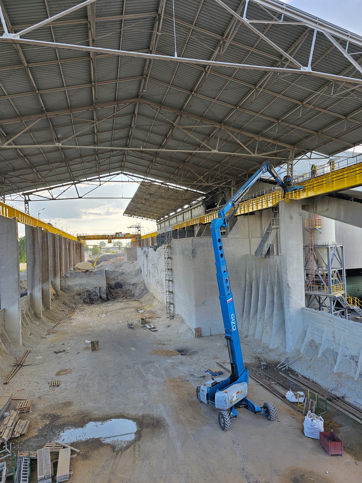 Wnętrze hali przemysłowej z podnośnikiem koszowym na tle betonowej konstrukcji i stalowej platformy z pracownikami.