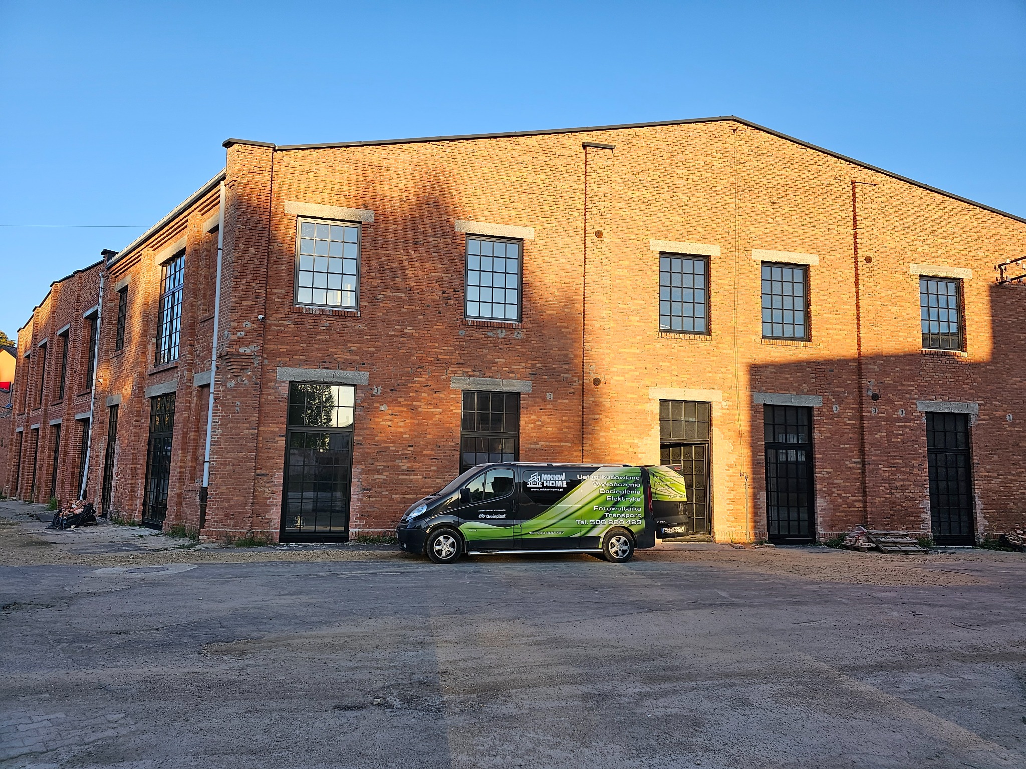 Ceglany budynek z czarnymi oknami i firmowy van z reklamą usług budowlanych na tle placu.