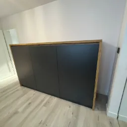 Nowoczesna szafa z trzema ciemnoszarymi frontami, obramowana drewnianym blatem i bokami, ustawiona na jasnej podłodze w minimalistycznym wnętrzu z białymi ścianami i drzwiami.