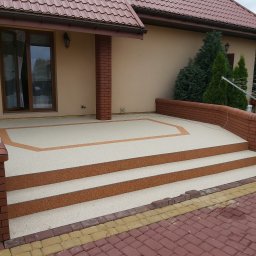 Roksy And Stonsy - Wykończony taras z beżową nawierzchnią żywiczną i brązowymi obramowaniami, prowadzący schodami do domu z czerwoną dachówką, widok z poziomu kostki brukowej.