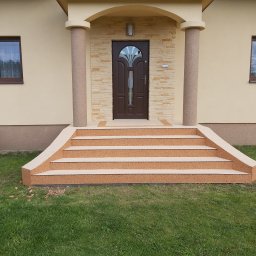 Roksy And Stonsy - Wejście do domu z czterostopniowymi schodami pokrytymi beżową nawierzchnią żywiczną, kolumny z tej samej nawierzchni podtrzymujące zadaszenie, ściany domu w kolorze kremowym, trawnik przed schodami.