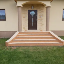 Wejście do domu z czterostopniowymi schodami pokrytymi beżową nawierzchnią żywiczną, kolumny z tej samej nawierzchni podtrzymujące zadaszenie, ściany domu w kolorze kremowym, trawnik przed schodami.