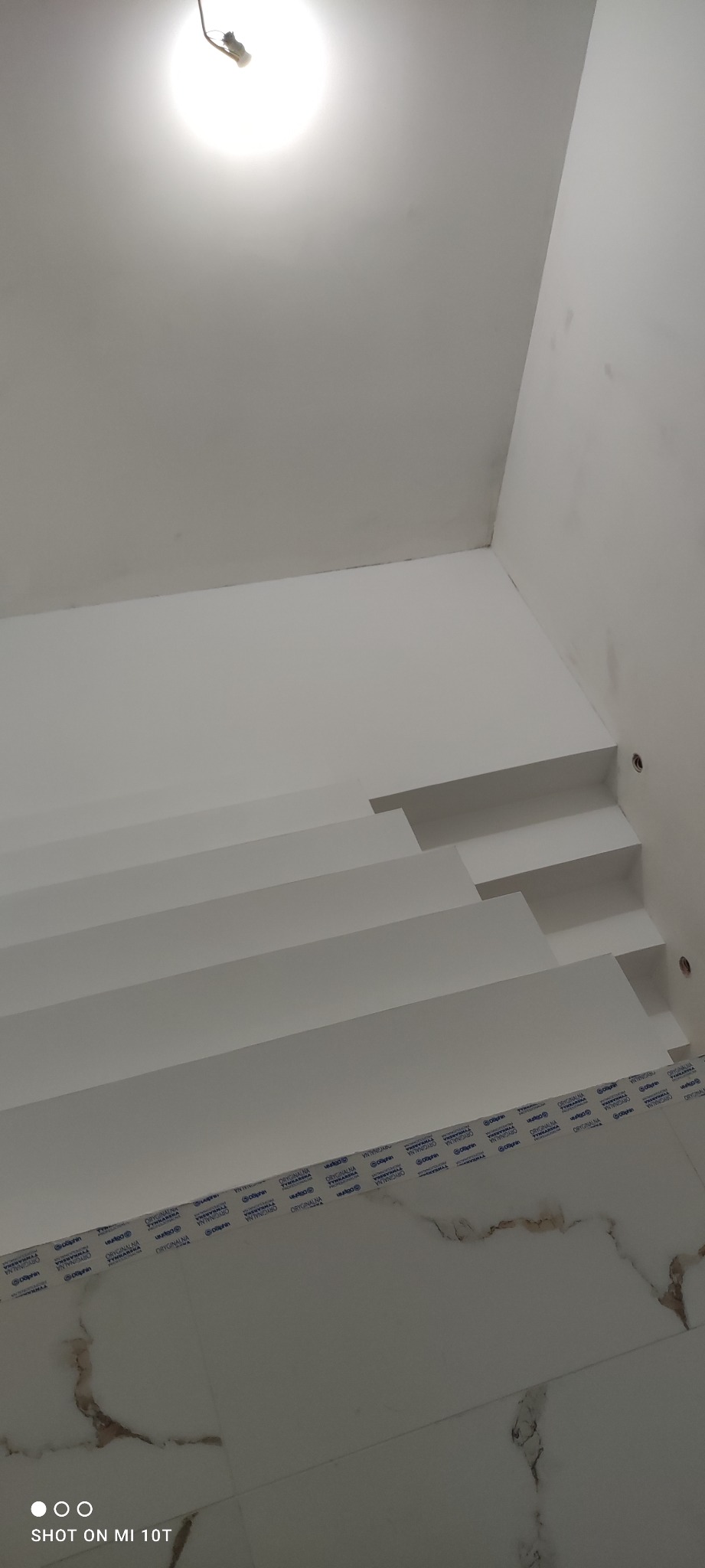 Nowoczesne schody wewnętrzne z białymi stopniami i marmurową posadzką, widok od dołu, z widocznym oświetleniem sufitowym.