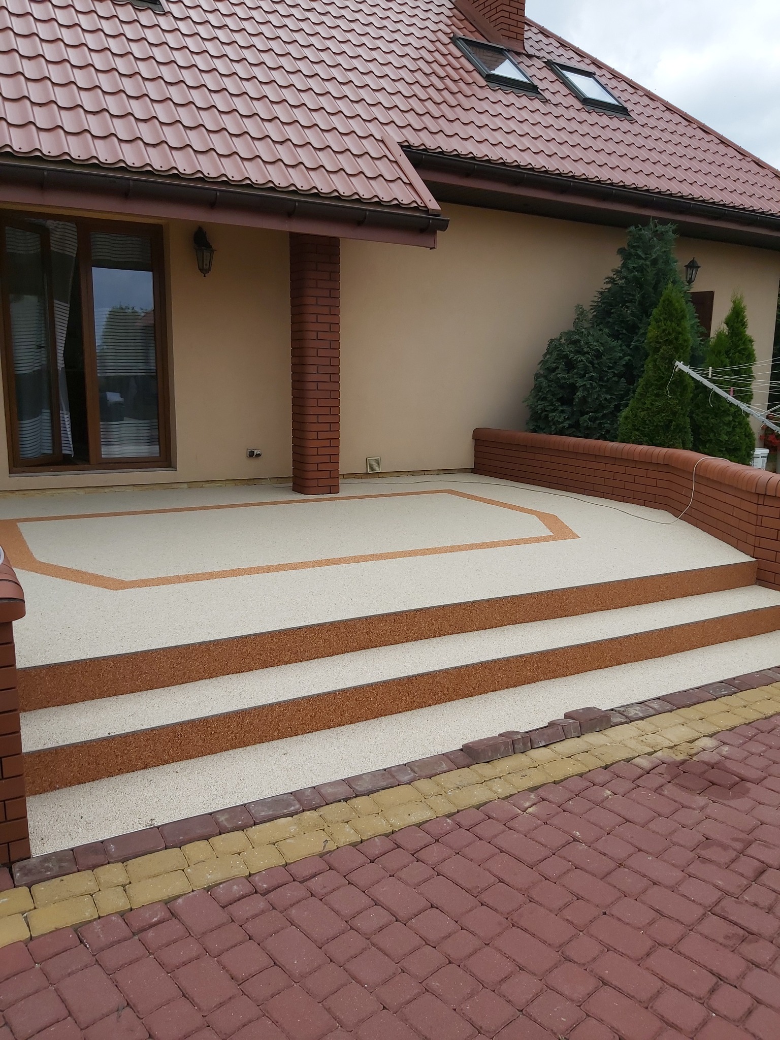 Wykończony taras z beżową nawierzchnią żywiczną i brązowymi obramowaniami, prowadzący schodami do domu z czerwoną dachówką, widok z poziomu kostki brukowej.