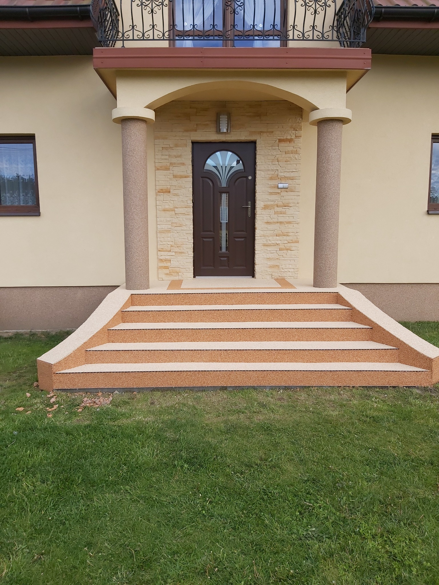 Wejście do domu z czterostopniowymi schodami pokrytymi beżową nawierzchnią żywiczną, kolumny z tej samej nawierzchni podtrzymujące zadaszenie, ściany domu w kolorze kremowym, trawnik przed schodami.