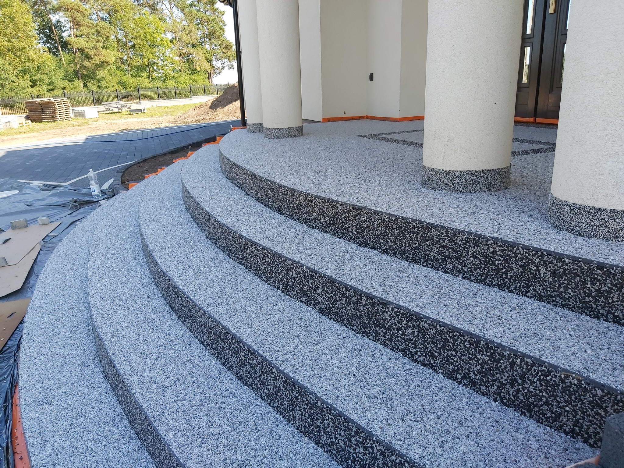 Zaokrąglone schody wejściowe z szarej posadzki kamyczkowej, wykończone ciemnoszarym obrzeżem, prowadzące do budynku z białymi kolumnami.