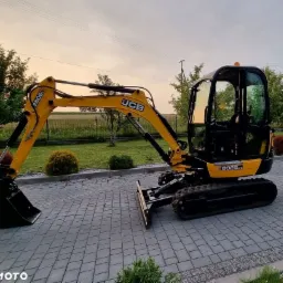 Żółto-czarna mini koparka JCB 8026 CTS na kostce brukowej, widok z boku, oświetlona popołudniowym słońcem.