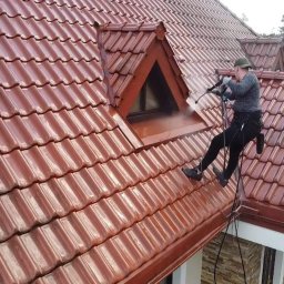 ProExtreme Tomasz Szempliński - Pracownik w uprzęży, myjący ciśnieniowo okno dachowe na czerwonym, błyszczącym dachu z dachówki ceramicznej, widok z dołu.