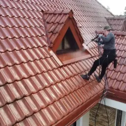 Pracownik w uprzęży, myjący ciśnieniowo okno dachowe na czerwonym, błyszczącym dachu z dachówki ceramicznej, widok z dołu.
